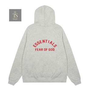 Op de afbeelding: Een lichtgrijze gem&ecirc;leerde hoodie met lange mouwen. Op de achterkant staat de rode tekst ESSENTIALS in een boog boven de rode tekst FEAR OF GOD. Het kledingstuk is voorzien van een geribbelde zoom en geribbelde mouwboorden.