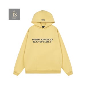 K&ouml;nnte beinhalten: Hellgelber Kapuzenpullover mit dem schwarzen Schriftzug "FEAR OF GOD essentials" auf der Brust. Der Kapuzenpullover hat eine Kapuze und ein kleines schwarzes Rechteck in der N&auml;he der Vorderseite der Kapuze. Die &Auml;rmel sind lang und das Material scheint weich zu sein.