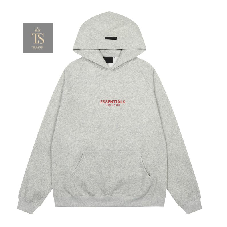 Op de afbeelding: Een lichtgrijze gem&ecirc;leerde hoodie met een capuchon voorzien van een zwarte rechthoekige patch. Op de borst staat de rode tekst ESSENTIALS FEAR OF GOD. Het kledingstuk heeft lange mouwen, een kangoeroezak en boorden aan de mouwen en zoom.