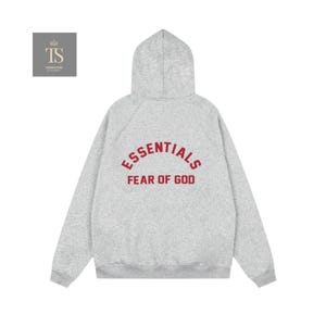 Może przedstawiać: Jasnoszara bluza z kapturem z kapturem i długimi rękawami. Z tyłu bluzy znajduje się napis "ESSENTIALS" wygięty nad "FEAR OF GOD" w czerwonej, łukowatej czcionce. Bluza wykonana jest z miękkiego, wygodnego materiału.