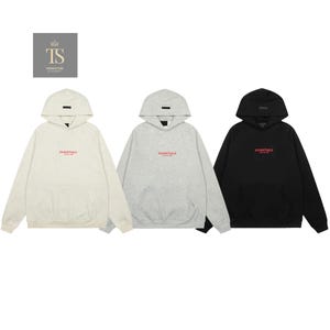 Op de afbeelding: Drie hoodies met lange mouwen in crème, lichtgrijs en zwart. Elke hoodie heeft een kangoeroezak, een capuchon met een kleine rechthoekige patch en de tekst ESSENTIALS FEAR OF GOD in het rood op de borst gedrukt.