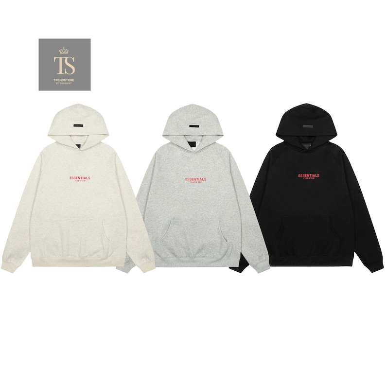 Op de afbeelding: Drie hoodies met lange mouwen in cr&egrave;me, lichtgrijs en zwart. Elke hoodie heeft een kangoeroezak, een capuchon met een kleine rechthoekige patch en de tekst ESSENTIALS FEAR OF GOD in het rood op de borst gedrukt.