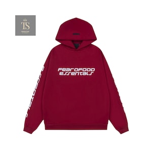 K&ouml;nnte beinhalten: Ein weinroter Kapuzenpullover mit dem wei&szlig;en Text "FEAR OF GOD essentials" auf der Brust. Die &Auml;rmel haben ebenfalls wei&szlig;en Text. Der Hoodie hat eine Kapuze und ein kleines schwarzes Etikett auf der Kapuze.
