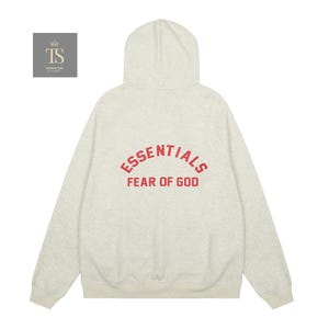 Op de afbeelding: Een lichtgrijs gem&ecirc;leerde hoodie met lange mouwen, een capuchon, geribbelde boorden en een geribbelde zoom. Op de achterkant staat de rode tekst ESSENTIALS in een boog boven de tekst FEAR OF GOD. Een klein vierkant logo met de letters TS is zichtbaar.