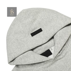 Op de afbeelding: Een lichtgrijze gem&ecirc;leerde hoodie met een opvallende zwarte rechthoekige patch in het midden van de capuchon. De binnenkraag toont een wit label met maat M en een zwart merklabel dat onder de opening van de capuchon is geplaatst.