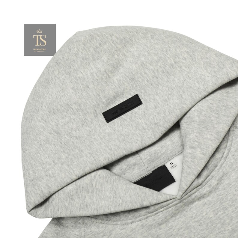 Op de afbeelding: Een lichtgrijze gem&ecirc;leerde hoodie met een opvallende zwarte rechthoekige patch in het midden van de capuchon. De binnenkraag toont een wit label met maat M en een zwart merklabel dat onder de opening van de capuchon is geplaatst.