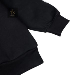 K&ouml;nnte beinhalten: Schwarzes Sweatshirt mit gerippten B&uuml;ndchen und Saum. Der &Auml;rmel hat ein wei&szlig;es Design am oberen Teil. Das Logo "TS TRENDSTORE" befindet sich in einem schwarzen Feld mit einer goldenen Krone.