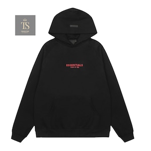 Op de afbeelding: Een zwarte hoodie met lange mouwen, een kangoeroezak aan de voorzijde en een capuchon. Op de borst staat de rode tekst ESSENTIALS FEAR OF GOD. Links bovenaan bevindt zich een klein vierkant logo met de letters TS en een kroonicoon.