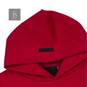 K&ouml;nnte beinhalten: Nahaufnahme eines roten Hoodies mit Kapuze. Der Hoodie hat ein kleines schwarzes rechteckiges Etikett in der N&auml;he der Kapuze. Das Logo "TS TRENDSTORE" befindet sich in der oberen linken Ecke.