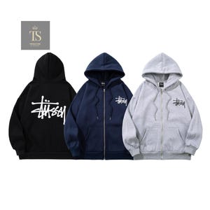 Può includere: Tre felpe con cappuccio in nero, blu navy e grigio melange. La felpa nera presenta un grande logo Stüssy bianco sul davanti, mentre le felpe con zip blu navy e grigie mostrano un logo Stüssy nero più piccolo sul lato sinistro del petto.