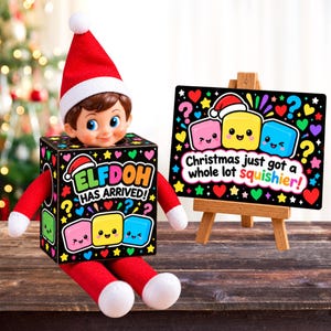 Könnte beinhalten: Eine Puppe mit rot-weißem Hut sitzt in einer schwarzen Schachtel mit der Aufschrift "ELFDOH HAS ARRIVED!" und bunten Herzen und Sternen. Daneben steht eine kleine Holzstaffelei mit einem Schild "Christmas just got a whole lot squishier!" und drei lächelnden, bunten quadratischen Figuren.