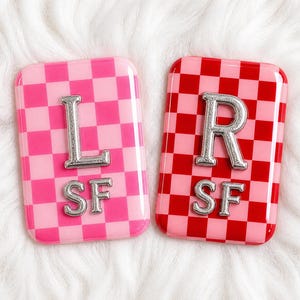 Op de afbeelding: Twee rechthoekige, glanzende accessoires met een dambordpatroon. Eén is roze met een zilveren "L" en "SF", de andere is rood met een zilveren "R" en "SF". De items zijn waarschijnlijk decoratief of functioneel.