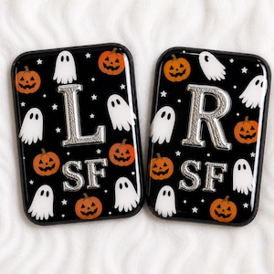 以下が含まれることがあります： ハロウィーンをテーマにした黒い携帯電話ケース2つ。オレンジ色のカボチャ、白い幽霊、銀色の文字が特徴です。片方のケースには文字「L」、もう片方には「R」があります。両方のケースに文字「SF」があります。