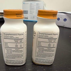 May include: Two white supplement bottles with orange caps. Each bottle has a label with "Supplement Facts" and ingredient information. The bottles are on a dark surface.
