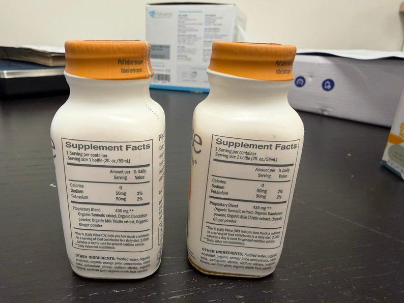 May include: Two white supplement bottles with orange caps. Each bottle has a label with "Supplement Facts" and ingredient information. The bottles are on a dark surface.