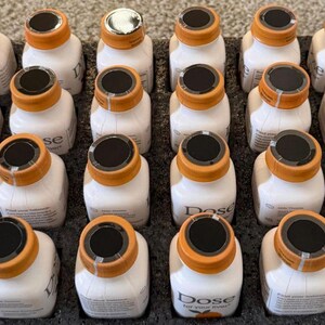 May include: A tray of small, white bottles with orange caps and black tops. The bottles are arranged in neat rows. The text "Dose" is visible on the bottles. The tray is black.