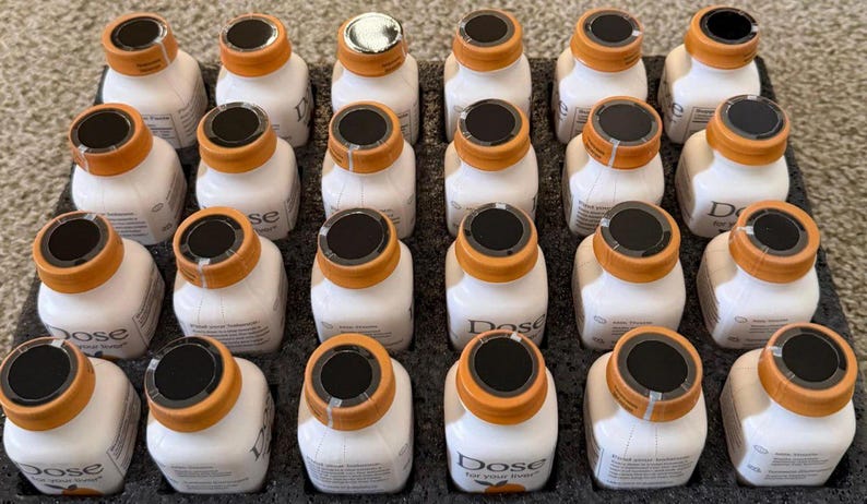 May include: A tray of small, white bottles with orange caps and black tops. The bottles are arranged in neat rows. The text "Dose" is visible on the bottles. The tray is black.