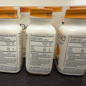 May include: White supplement bottles with orange caps are arranged in front of a box. Each bottle has a label with supplement facts, including calories, sodium, and potassium content. The bottles are filled with a liquid and are ready to drink.