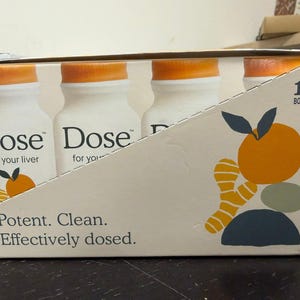 May include: A box containing 12 bottles of "Dose" drink. The bottles are white with orange caps. The box has text that reads "Potent. Clean. Effectively dosed." and a graphic of an orange and ginger.