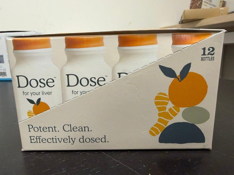 May include: A box containing 12 bottles of "Dose" drink. The bottles are white with orange caps. The box has text that reads "Potent. Clean. Effectively dosed." and a graphic of an orange and ginger.