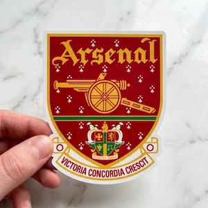 May include: A shield-shaped sticker featuring the Arsenal football club crest in red, gold, and green. The design includes a gold cannon, the text Arsenal, and the Latin motto Victoria Concordia Crescit, set against a red background with white symbols.