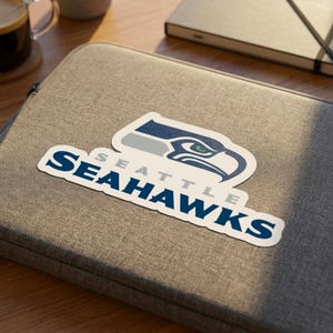 May include: A die-cut vinyl sticker featuring the Seattle Seahawks logo, which includes a stylized navy blue, grey, and green bird head graphic above the text SEATTLE SEAHAWKS in bold navy blue lettering with a white border on a grey laptop sleeve.