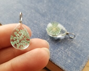 Op de afbeelding: Twee traanvormige glazen hangers met zilveren draadlussen. Elke hanger bevat een geperste groene bloem.