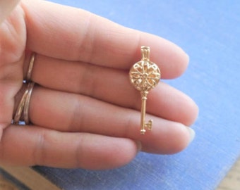 May include: A gold-toned metal key pendant featuring a circular head with an intricate, openwork floral pattern and a slender shaft ending in a small, simple bit. The key has a polished finish and a small loop at the top for attachment.