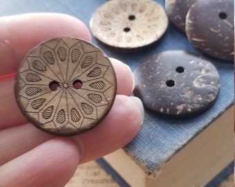 以下が含まれることがあります： 2つの穴のある茶色のココナッツの殻のボタンのコレクション。ボタンの一部には彫刻が施され、一部はプレーンです。ボタンは青い本の上に置かれています。