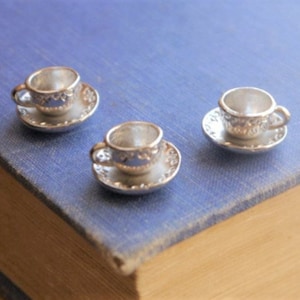 May include: Three miniature silver teacups and saucers with ornate designs. The cups and saucers are arranged on a blue book.