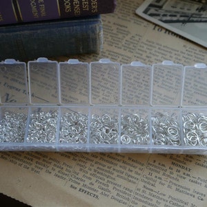 May include: A clear plastic storage box with 10 compartments filled with silver jump rings. The box is on a brown and white patterned surface.