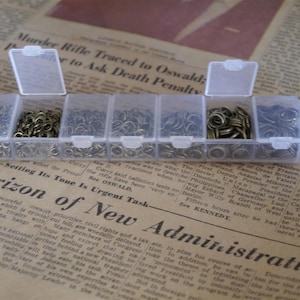 May include: A clear plastic storage box with six compartments, each filled with small, gold-coloured metal jump rings. The box is open and sitting on a newspaper.
