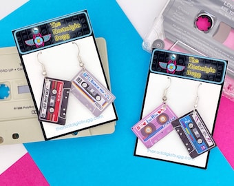 May include: Two pairs of cassette tape earrings. The earrings are silver with a black, red, and white cassette tape charm. The cassette tapes have the text "Hip Hop Mix", "Dance Mix", "Love Mix", and "Rock Mix" on them. The earrings are displayed on a white card with the text "The Nostalgia Bugg" on it.