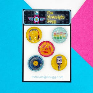 May include: Set of six colourful round stickers with a retro music theme. The stickers feature a pair of headphones, a record, a microphone, a graphic equaliser, and a stylised record player. The stickers are on a white background with the text "The Nostalgia Bugg" and "thenostalgiabugg.com".
