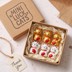 Peut inclure: Un ensemble de six figurines de chats porte-bonheur miniatures dans une boîte en carton marron marquée MINI LUCK CATS. Trois chats sont dorés avec des détails rouges, et trois sont blancs avec des détails rouges. Une étiquette Thank You est attachée.