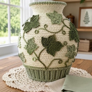May include: A cream-colored crochet vase with green ivy leaf accents. The vase has a wide base and a flared top, with detailed leaf and vine designs. It sits on a crocheted doily, with a folded napkin nearby. The vase is a decorative home accent.