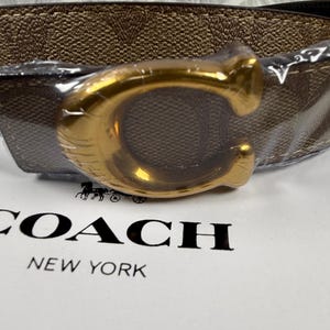 Könnte beinhalten: Wendegürtel in Braun und Schwarz mit goldfarbener Schnalle in Form des Buchstabens "C". Der Gürtel ist in klarem Kunststoff verpackt. Die Wörter "COACH" und "NEW YORK" sind auf der weißen Oberfläche aufgedruckt.
