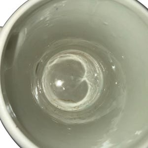 May include: A top-down view of a white ceramic mug with a smooth, glossy interior finish. The cylindrical vessel features a plain, unadorned white surface inside and along the rim, showing a slight depth and a circular base at the bottom.