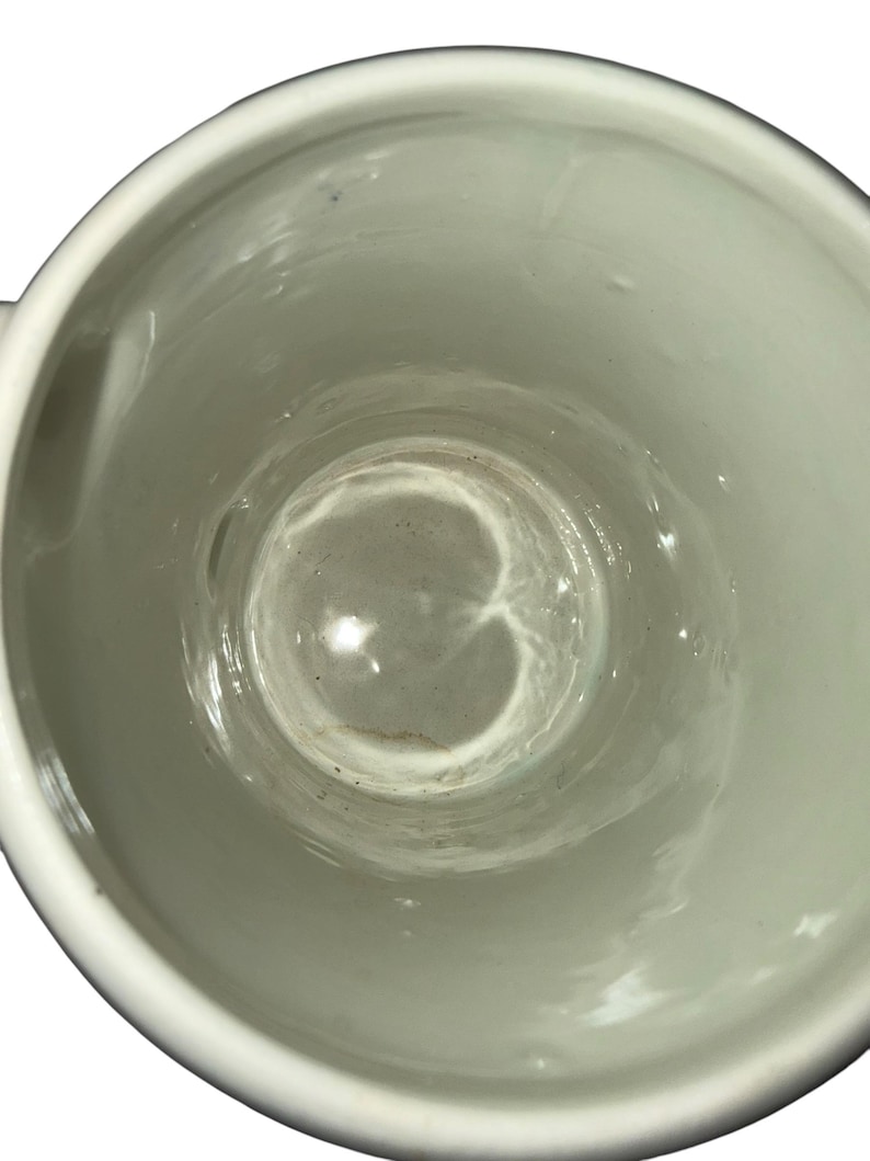 May include: A top-down view of a white ceramic mug with a smooth, glossy interior finish. The cylindrical vessel features a plain, unadorned white surface inside and along the rim, showing a slight depth and a circular base at the bottom.