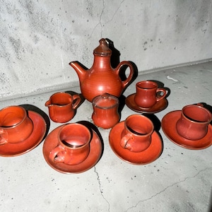May include: A terracotta-coloured tea set, including a teapot, creamer, sugar bowl, and six cups with saucers. The set is made of ceramic and has a rustic, handmade appearance. The cups and saucers are approximately 7.6 cm in diameter.