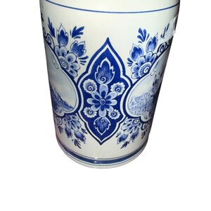 May include: A cylindrical ceramic beer stein featuring a white base with intricate blue floral patterns and detailed landscape illustrations. The vessel has a smooth finish, a wide rim, and a decorative central motif surrounded by foliage.