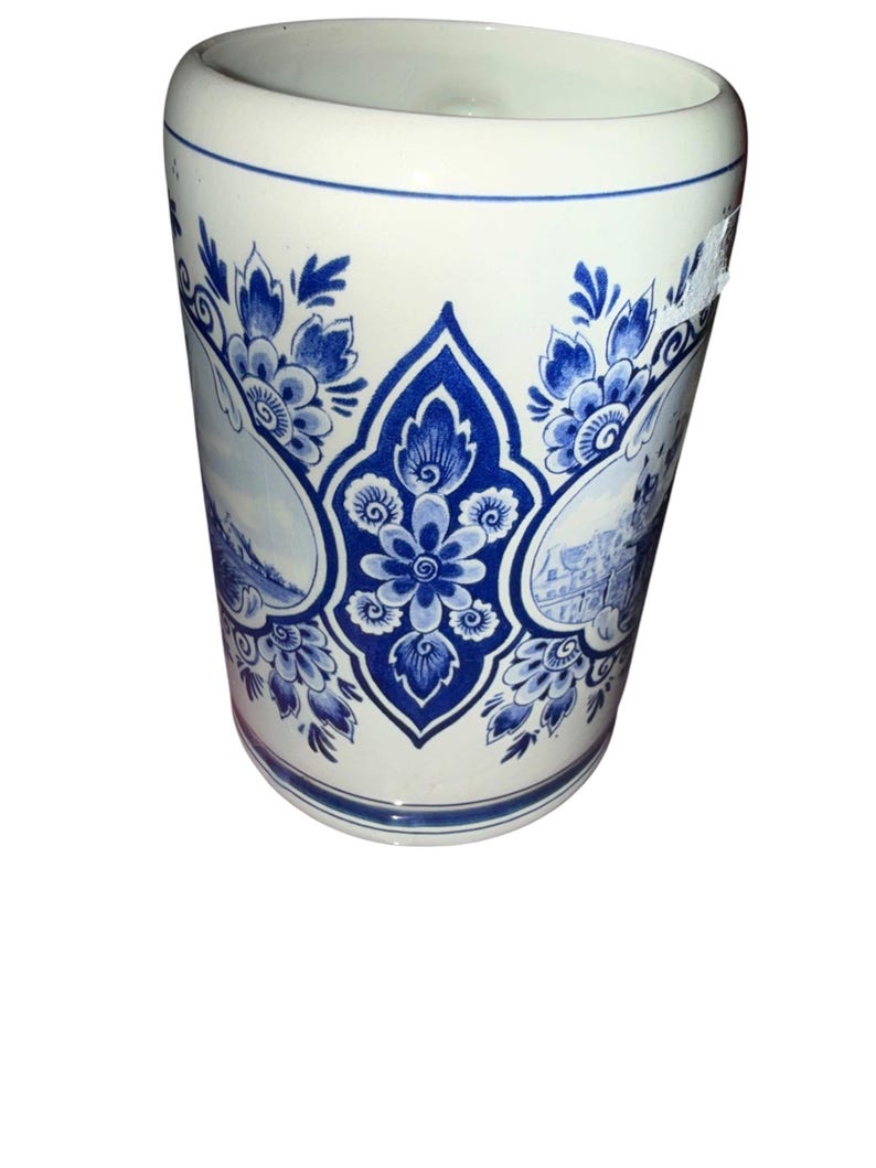May include: A cylindrical ceramic beer stein featuring a white base with intricate blue floral patterns and detailed landscape illustrations. The vessel has a smooth finish, a wide rim, and a decorative central motif surrounded by foliage.