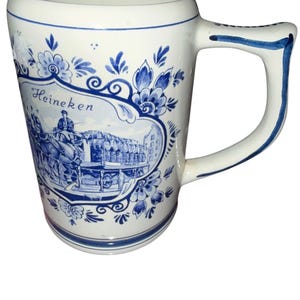May include: A white ceramic beer mug featuring blue floral patterns and a central illustration of a horse-drawn carriage carrying barrels. The text Heineken is printed above the carriage scene. The mug has a sturdy handle with blue trim.