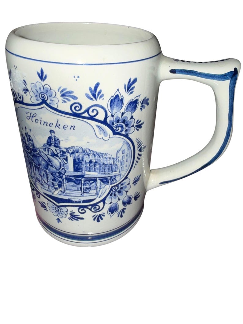 May include: A white ceramic beer mug featuring blue floral patterns and a central illustration of a horse-drawn carriage carrying barrels. The text Heineken is printed above the carriage scene. The mug has a sturdy handle with blue trim.