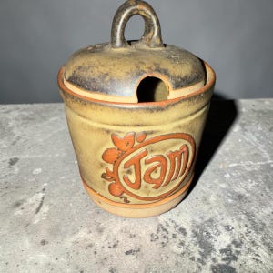 Puede incluir: Un tarro de cerámica color canela para mermelada con tapa a juego que incluye un asa de lazo y una muesca para cuchara. El frente del tarro muestra la palabra Jam en letras naranjas oscuras en relieve con un diseño floral grabado.