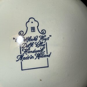 May include: A white ceramic base featuring a blue printed stamp. The stamp displays a stylized house icon above the text "'t Delftsche Huys" followed by "Delft Blue", "Handwork", and "Made in Holland" enclosed within a rectangular border.