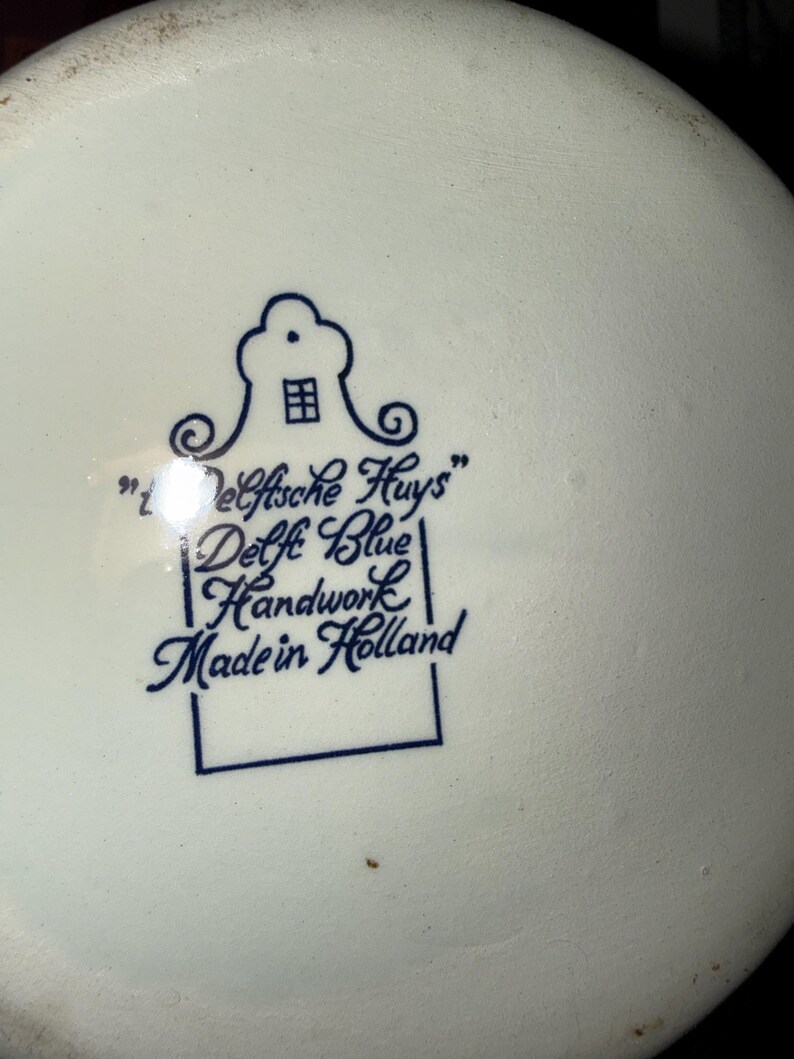 May include: A white ceramic base featuring a blue printed stamp. The stamp displays a stylized house icon above the text "'t Delftsche Huys" followed by "Delft Blue", "Handwork", and "Made in Holland" enclosed within a rectangular border.