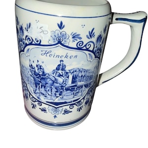 May include: A white ceramic beer mug featuring blue floral patterns and a central illustration of a horse-drawn carriage carrying barrels. The text Heineken is printed in blue script above the carriage. A small white sticker with 50 is attached.
