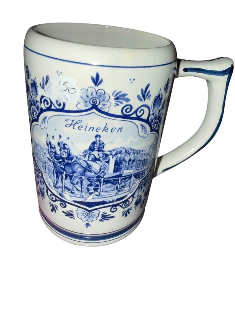 May include: A white ceramic beer mug featuring blue floral patterns and a central illustration of a horse-drawn carriage carrying barrels. The text Heineken is printed in blue script above the carriage. A small white sticker with 50 is attached.