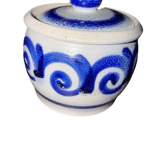 May include: A small, ceramic jar with a lid. The jar is white with blue swirl designs and a blue knob on the lid. The jar has a round shape and a slightly textured surface.